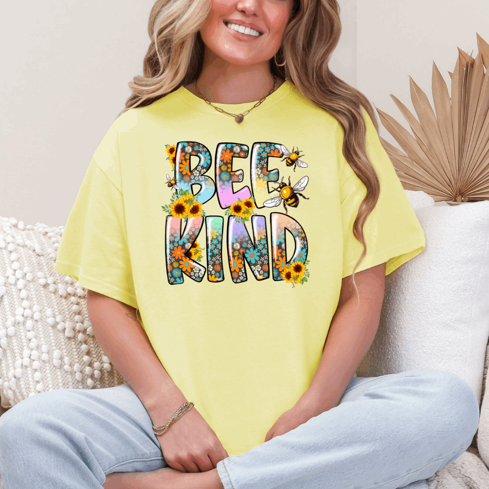 Woman wearing a yellow Bee Kind T-shirt with colorful floral and bee design, sitting cross-legged and smiling.