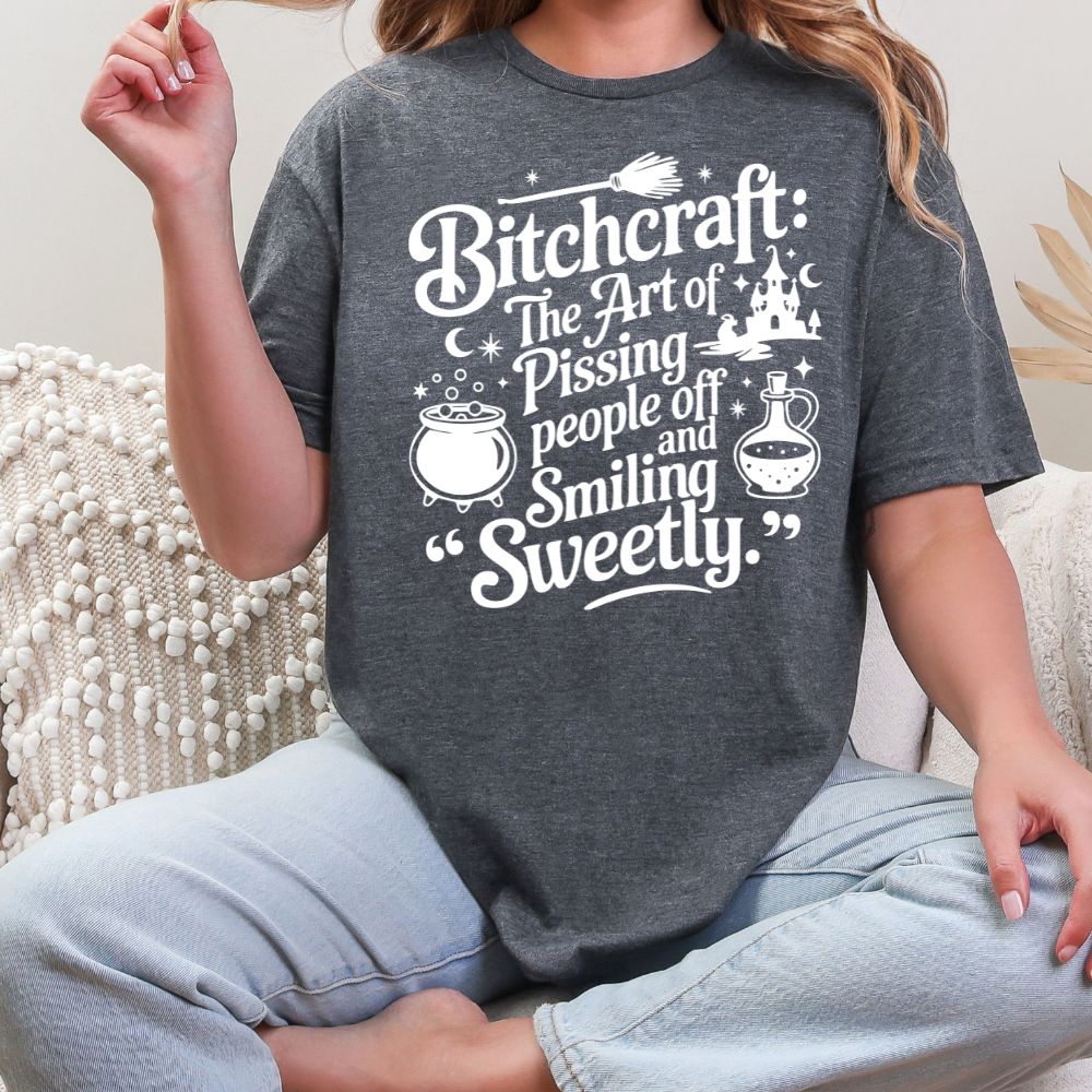 Dark heather T-shirt with white graphic saying “Bitchcraft: The art of pissing people off and smiling sweetly” with witchy icons