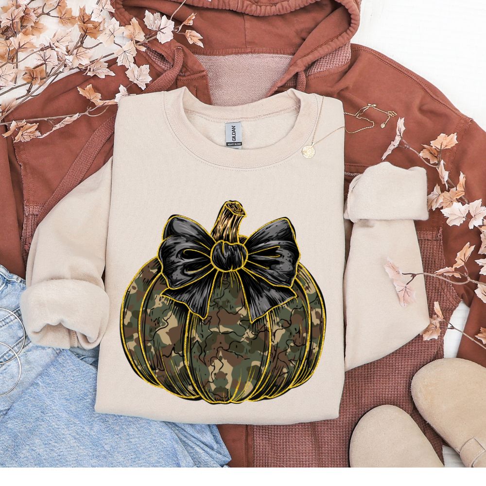 Beige sweatshirt with a camouflage pumpkin and black bow design on a brown fabric background.