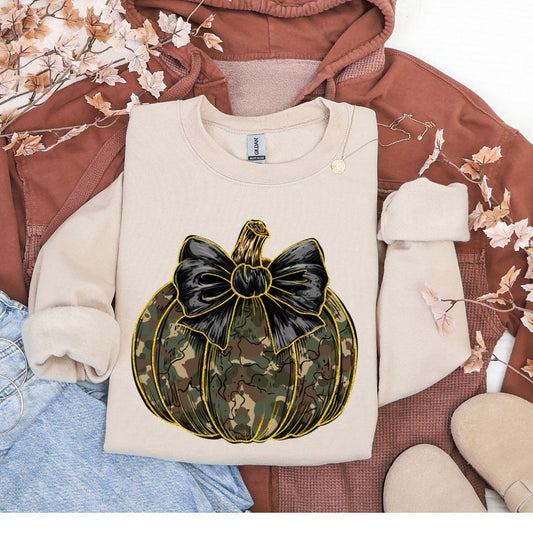 Beige sweatshirt with a camouflage pumpkin and black bow design on a brown fabric background.