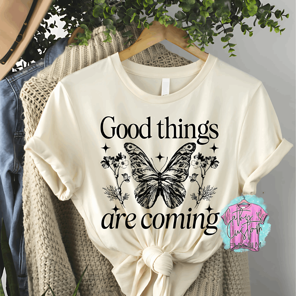 Beige T-shirt with "Good Things Are Coming" printed in black, featuring a detailed butterfly, floral accents, and celestial stars.