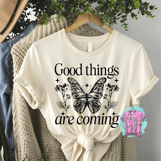 Beige T-shirt with "Good Things Are Coming" printed in black, featuring a detailed butterfly, floral accents, and celestial stars.