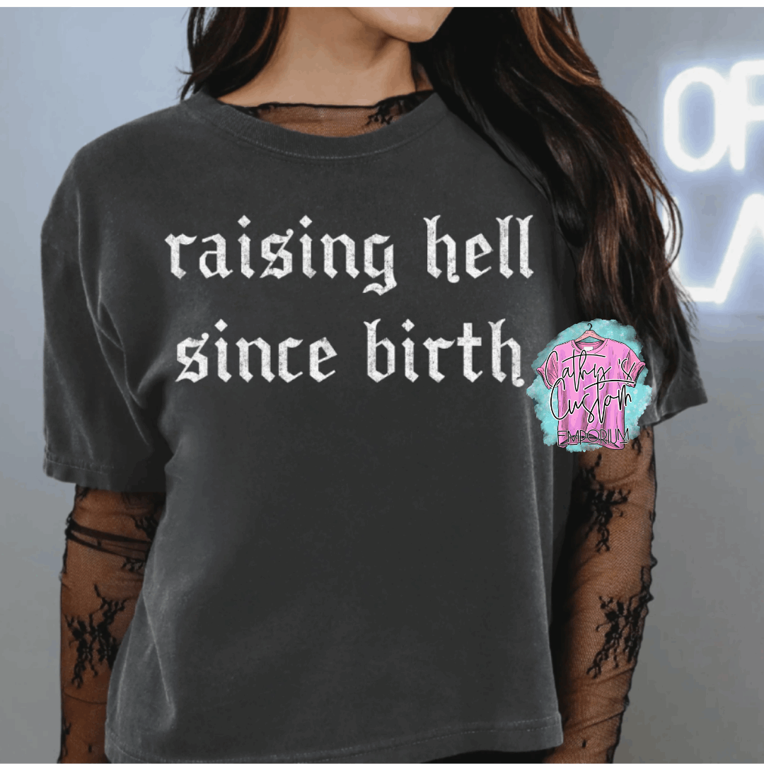 Black graphic T-shirt with bold white gothic text reading “raising hell since birth.” Styled for an edgy, rebellious look.