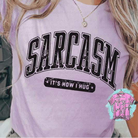 Pink Graphic T-shirt with "Sarcasm: It’s How I Hug" printed in bold, distressed varsity-style black font.