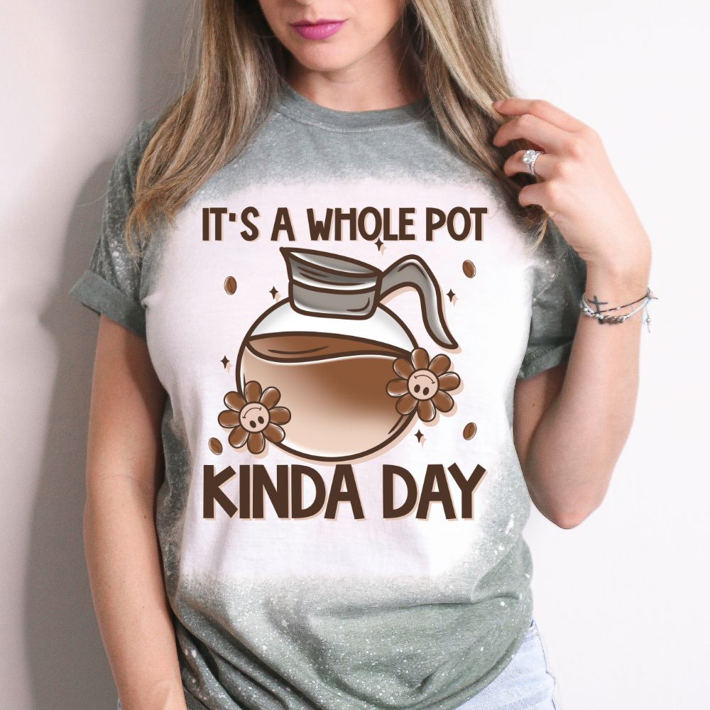 Woman wearing a green bleached T-shirt with a cartoon coffee pot and text that reads “It’s a Whole Pot Kinda Day,” surrounded by flowers and coffee beans.