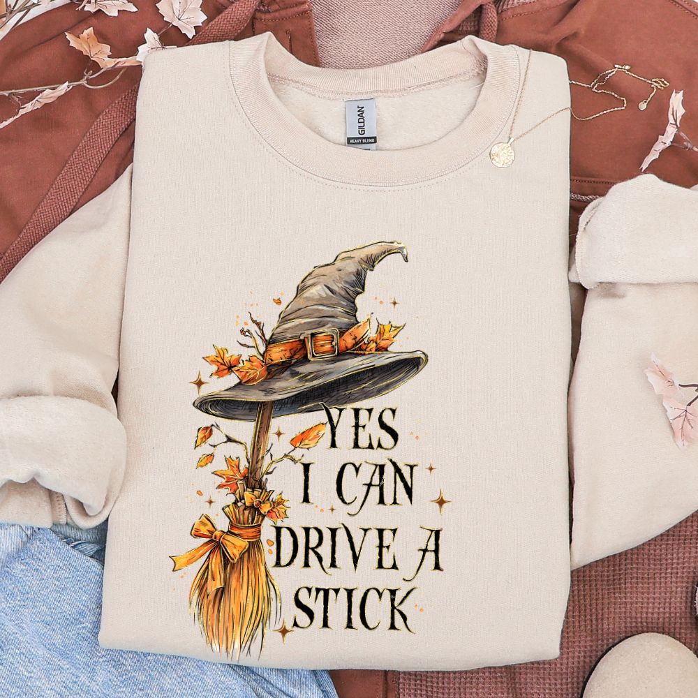 Beige sweatshirt with witch hat and broom design on a patterned surface. Yes I can drive a stick