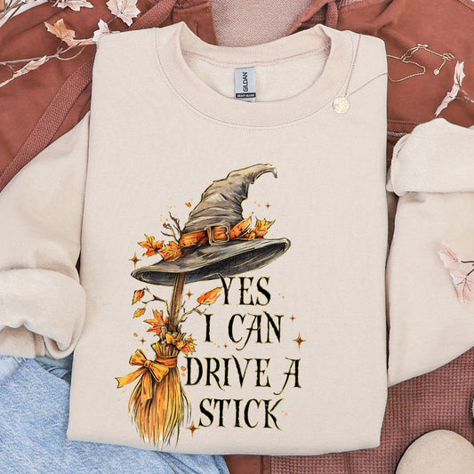 Beige sweatshirt with witch hat and broom design on a patterned surface. Yes I can drive a stick