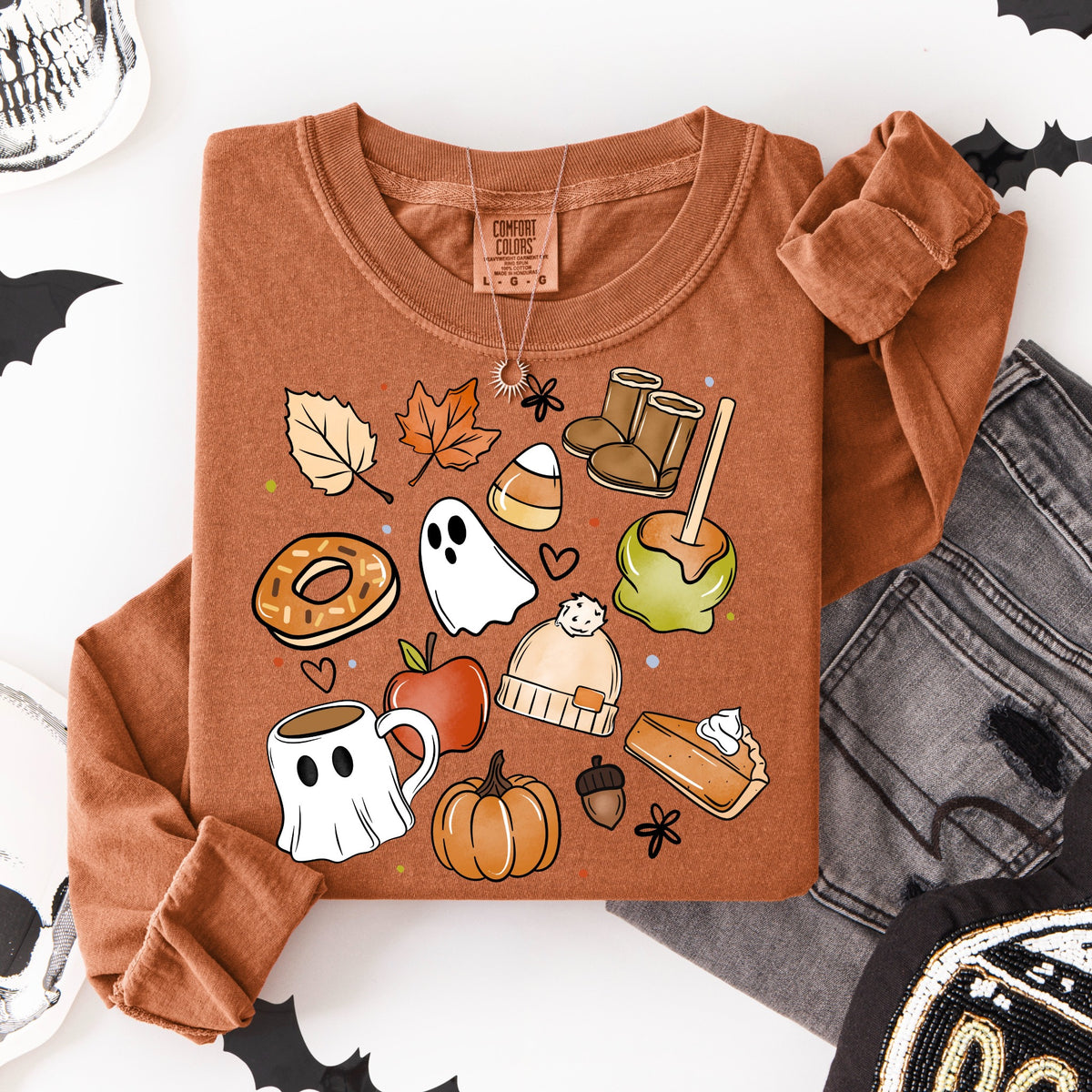 Flat lay of a rust-orange shirt featuring cozy fall favorites including a ghost, pumpkin, caramel apple, candy corn, coffee mug, doughnut, pie, boots, beanie, leaves, and acorn.