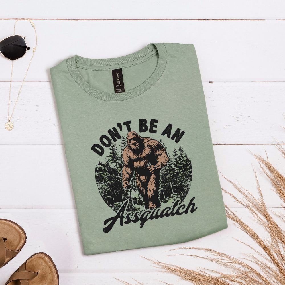 Sage green T-shirt with forest graphic and “Don’t be an Assquatch” phrase with Bigfoot in the center