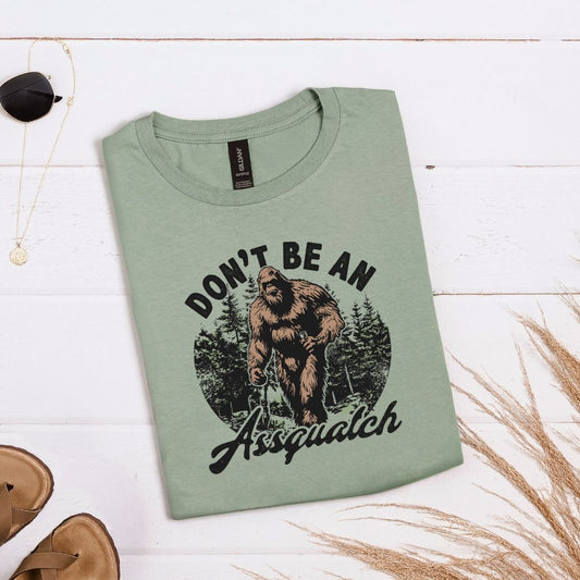 Sage green T-shirt with forest graphic and “Don’t be an Assquatch” phrase with Bigfoot in the center