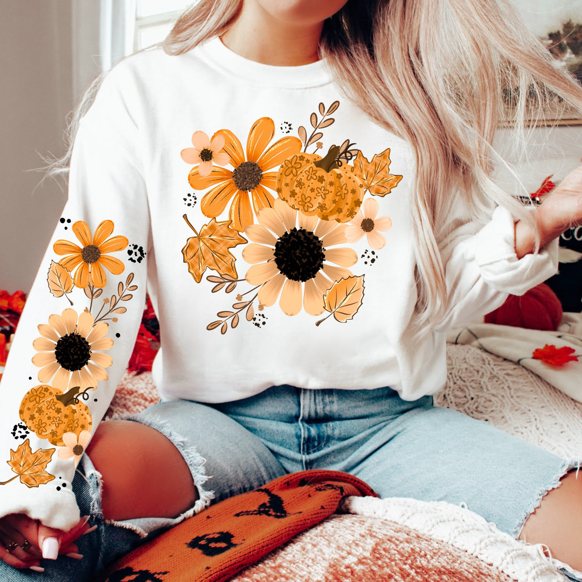 Woman wearing a white fall sweatshirt with orange and peach flowers, a floral pumpkin, autumn leaves, and coordinating sleeve designs