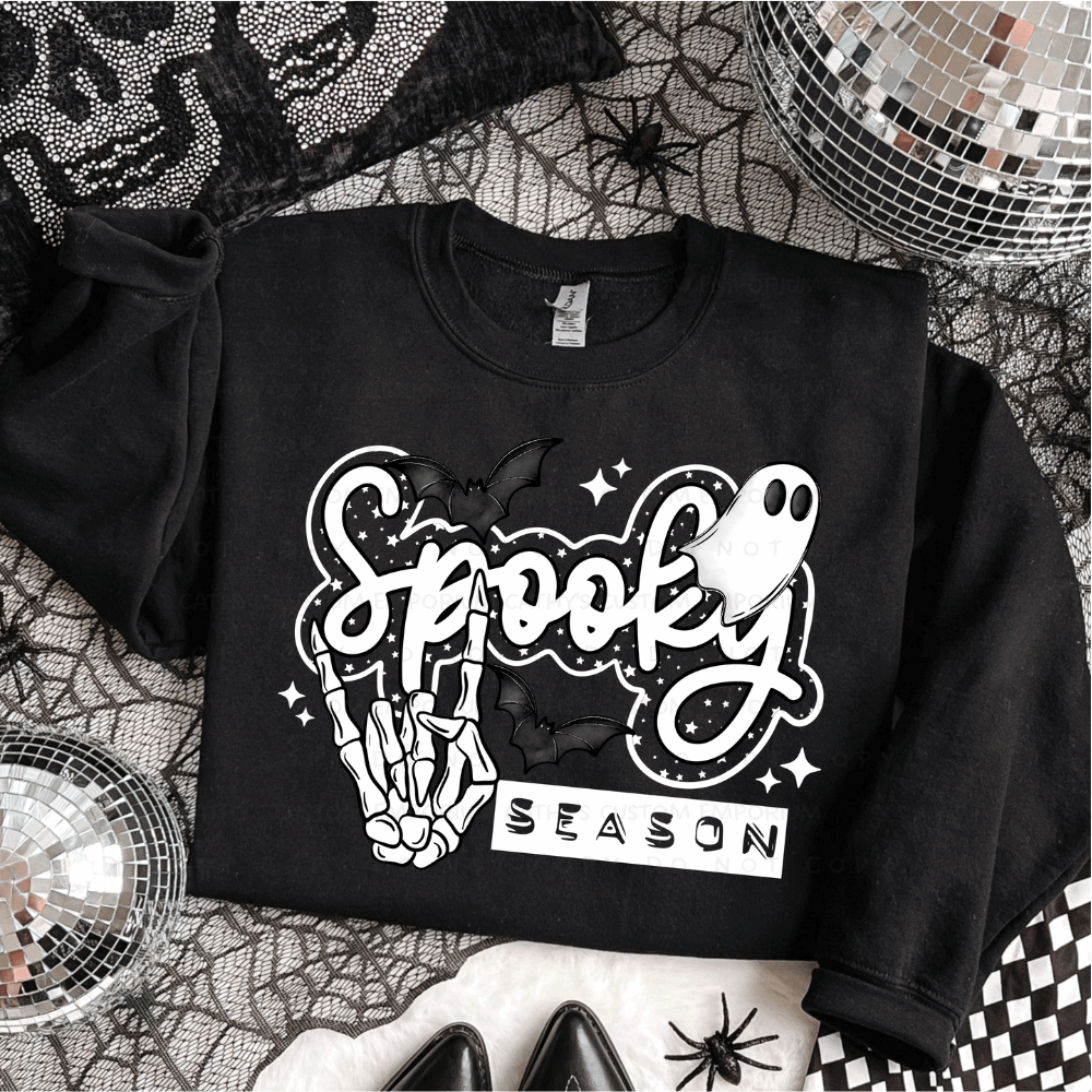 Black folded sweatshirt with a white Spooky Season Halloween graphic featuring a skeleton hand, ghost, bats, stars, and sparkles