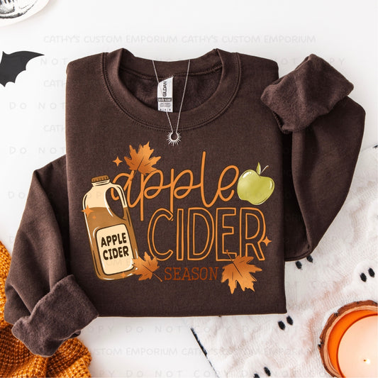 A folded brown crewneck sweatshirt with a 'Apple Cider Season' graphic, styled on a white background with autumn props including leaves, a candle, and a knit sweater.