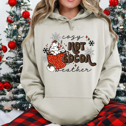 Person wearing a hoodie with 'cozy hot cocoa weather' design in front of a Christmas tree.