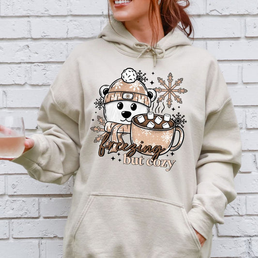 Person wearing a beige hoodie with a polar bear design and text Freezing, but Cozy.