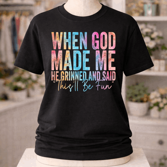 Black graphic T-shirt with colorful distressed text reading “When God Made Me He Grinned and Said This’ll Be Fun,” displayed on a mannequin.