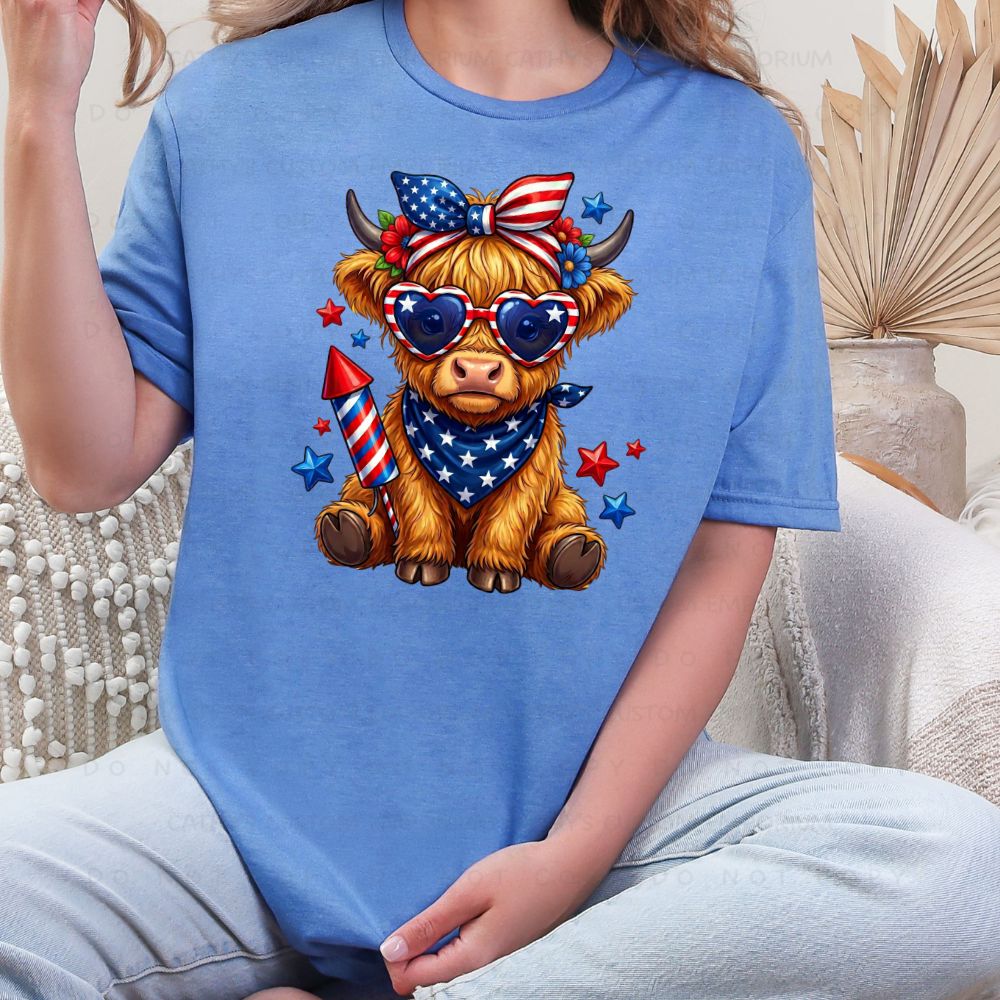 Person wearing a blue t-shirt with a cartoon Highland cow design featuring American flags and sunglasses.