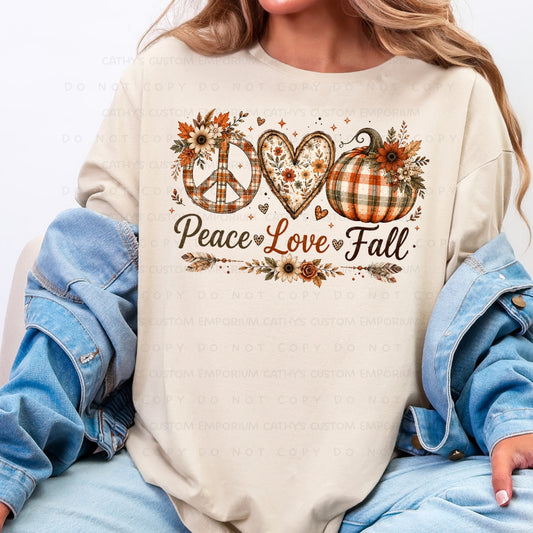 A woman wearing a cream-colored long-sleeve t-shirt with a fall-themed graphic that reads 'Peace Love Fall' in script, decorated with a peace sign, heart, pumpkin, and autumn leaves. She has a blue denim jacket draped off her shoulders and is wearing light-wash blue jeans.
