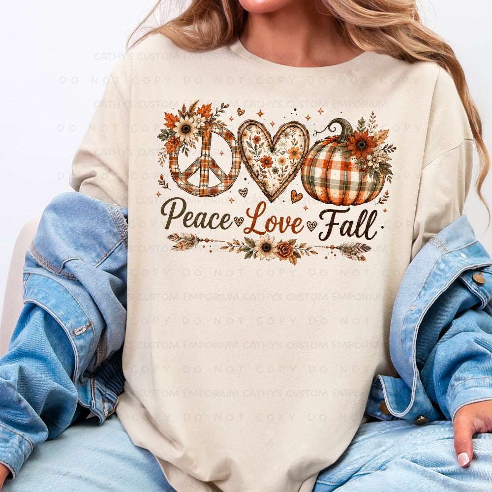 A woman wearing a cream-colored long-sleeve t-shirt with a fall-themed graphic that reads 'Peace Love Fall' in script, decorated with a peace sign, heart, pumpkin, and autumn leaves. She has a blue denim jacket draped off her shoulders and is wearing light-wash blue jeans.