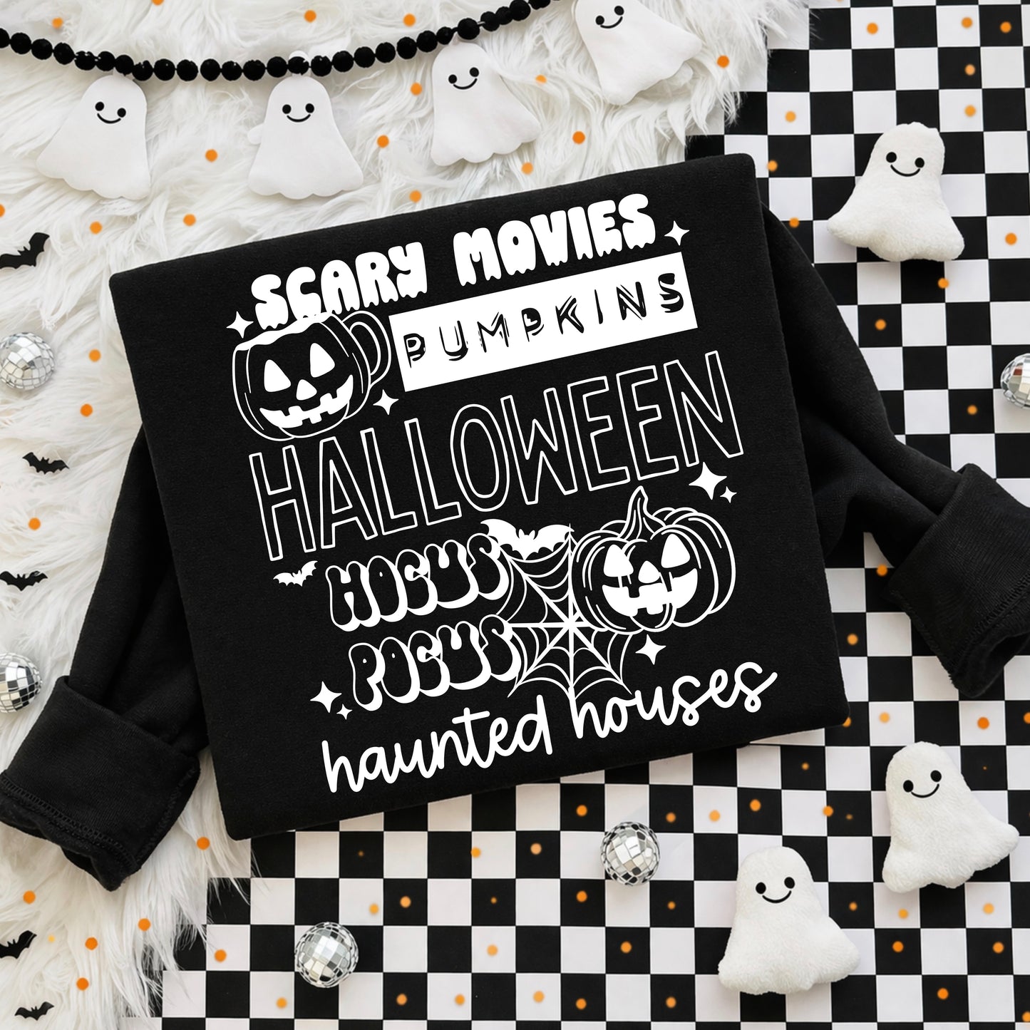 Flat lay of a black sweatshirt with a white stacked Halloween graphic featuring the words "Scary Movies," "Pumpkins," "Halloween," "Hocus Pocus," and "haunted houses" in retro fonts, with jack-o'-lanterns, spiderweb, and sparkle accents on a checkered and fur backdrop.