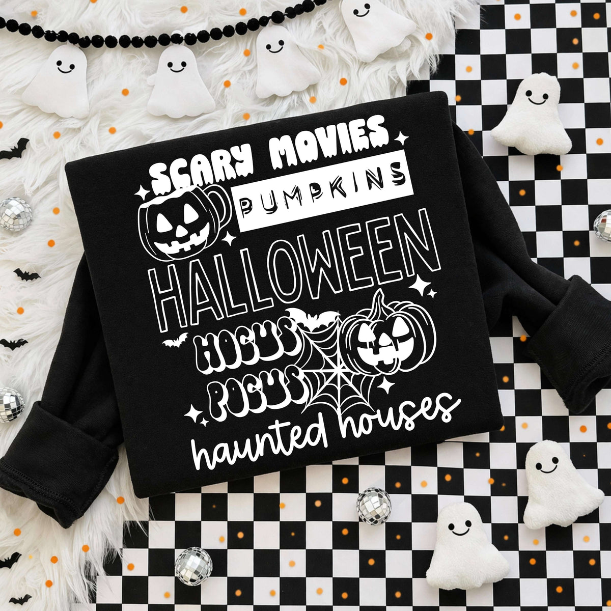 Flat lay of a black sweatshirt with a white stacked Halloween graphic featuring the words "Scary Movies," "Pumpkins," "Halloween," "Hocus Pocus," and "haunted houses" in retro fonts, with jack-o'-lanterns, spiderweb, and sparkle accents on a checkered and fur backdrop.