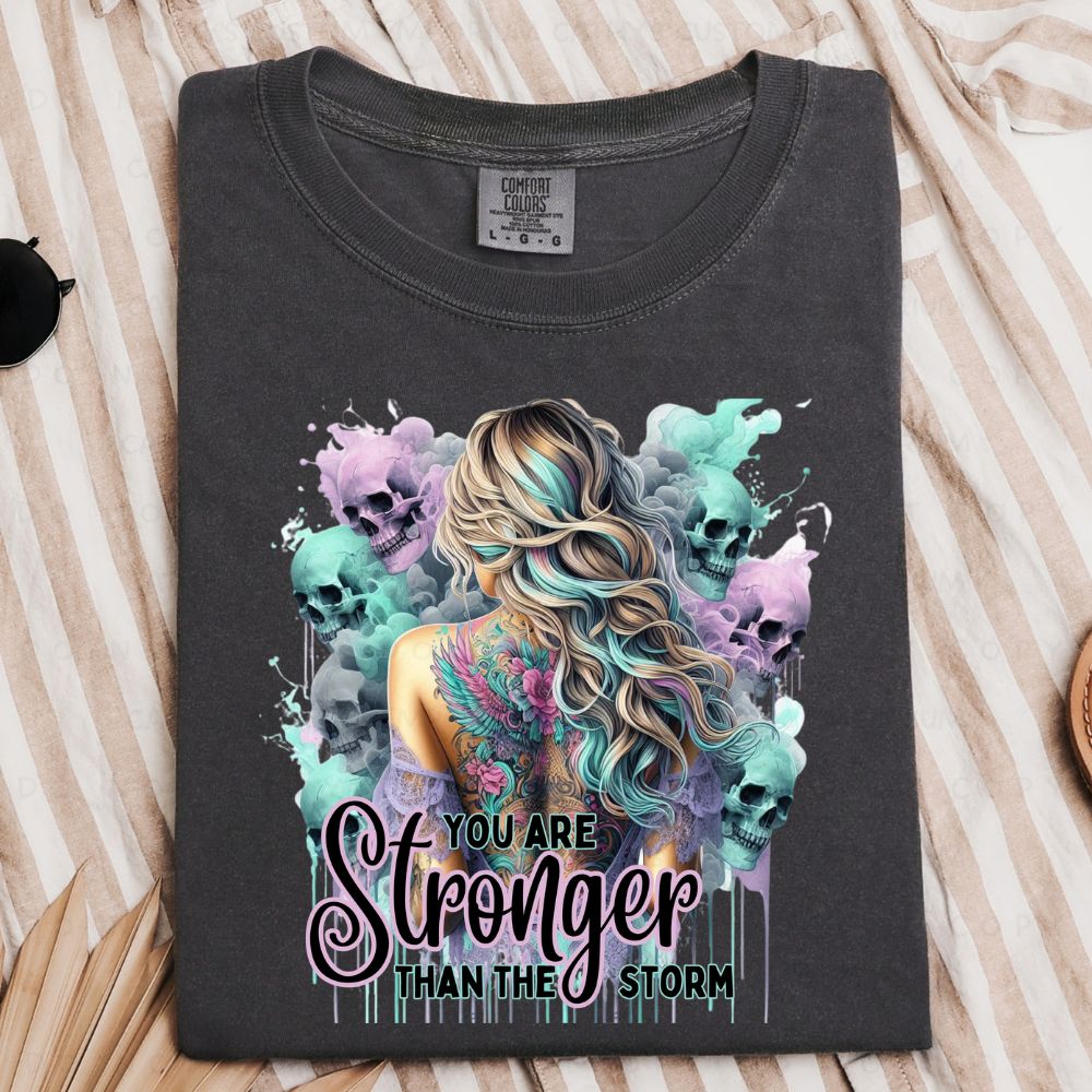 Black t-shirt with colorful graphic design and 'You are Stronger Than the Storm' text on a striped fabric background.