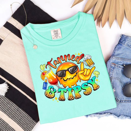 Mint green t-shirt with smiling sun in sunglasses, tropical drinks, and “Tanned & Tipsy” text.



