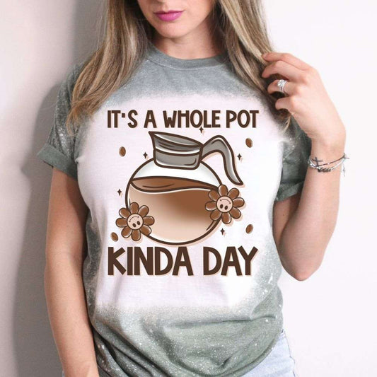 Woman wearing a green bleached T-shirt with a cartoon coffee pot and text that reads “It’s a Whole Pot Kinda Day,” surrounded by flowers and coffee beans.