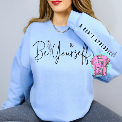 Light blue Be Yourself Graphic Sweatshirt with black cursive text, floral accents, and '& Don't Apologize' sleeve detail. Cozy unisex crewneck for confidence and self-expression