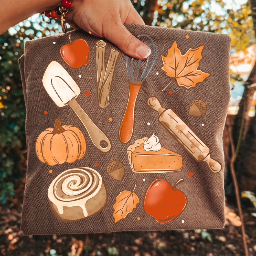 Folded brown fall graphic T-shirt featuring apples, pumpkin, pumpkin pie, cinnamon roll, rolling pin, whisk, spatula, acorns, and autumn leaves.