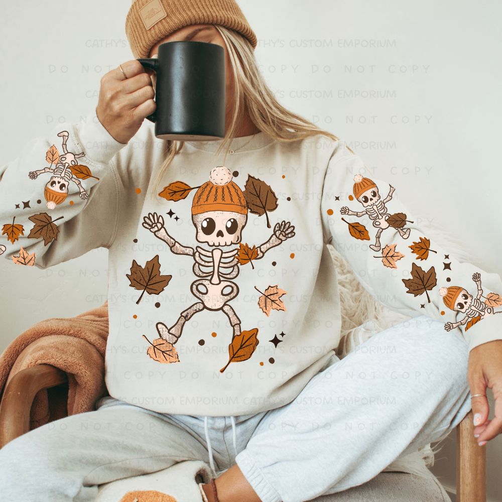 Woman in a tan knit beanie wearing a cream sweatshirt with playful skeletons in orange hats, falling autumn leaves, and coordinating sleeve designs