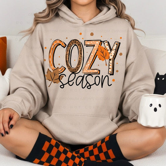 Woman wearing a sand-colored hoodie with a Cozy Season graphic in orange, tan, leopard print, and autumn leaf accents