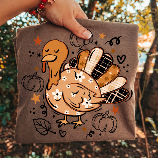 Folded brown Thanksgiving graphic T-shirt featuring a cute orange floral turkey with plaid feathers, pumpkins, leaves, stars, and hearts.