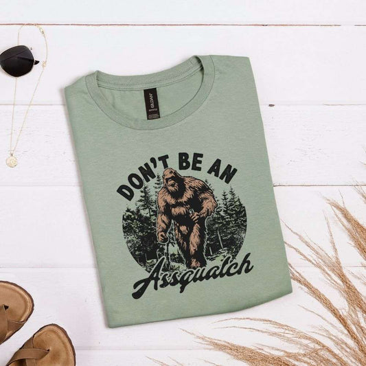 Sage green T-shirt with forest graphic and “Don’t be an Assquatch” phrase with Bigfoot in the center

