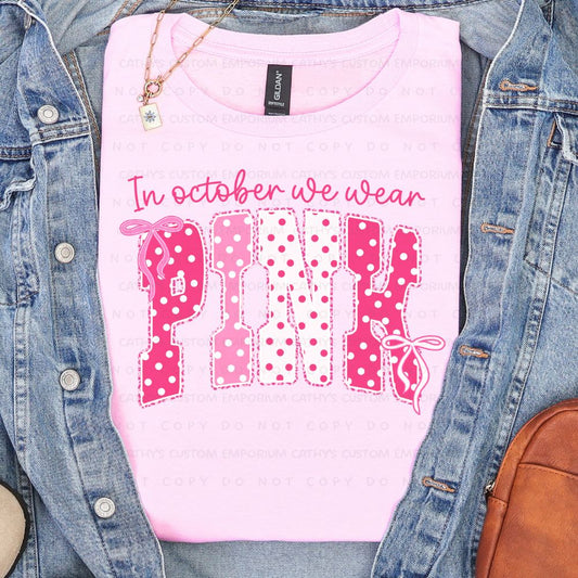 Light pink shirt featuring the phrase In October We Wear Pink in pink and white polka-dot lettering with bow accents.
