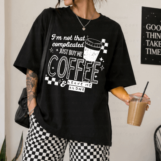 Black shirt featuring a white graphic that reads I'm Not That Complicated, Just Buy Me Coffee and Leave Me Alone with a coffee cup and checkerboard accents.