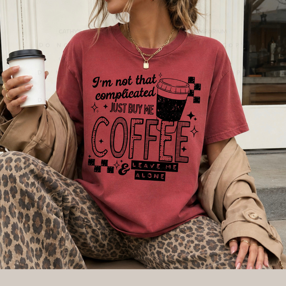 Muted red shirt featuring a black graphic that reads I'm Not That Complicated, Just Buy Me Coffee and Leave Me Alone with a coffee cup and checkerboard accents.