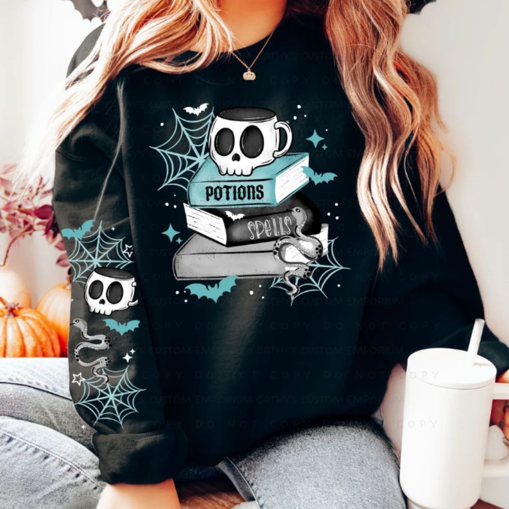 Woman wearing a black Halloween sweatshirt with stacked potion and spell books, a skull mug, snake, bats, spiderwebs, and coordinating sleeve artwork