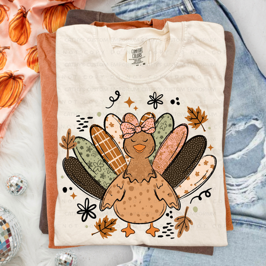 Folded cream Thanksgiving graphic T-shirt featuring a whimsical turkey with patterned floral feathers in orange, sage green, pink, cream, and brown.
