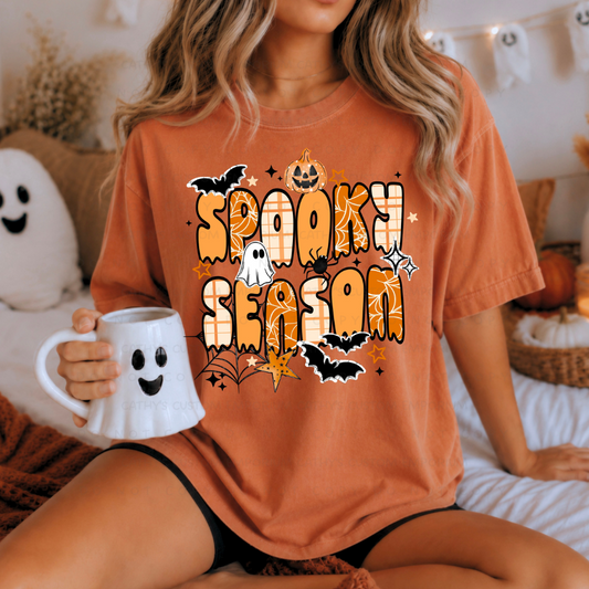 Woman wearing an orange Spooky Season Halloween graphic shirt with patterned retro lettering, ghosts, bats, a pumpkin, spiderwebs, and stars
