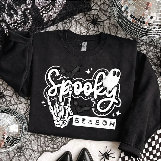 Black folded sweatshirt with a white Spooky Season Halloween graphic featuring a skeleton hand, ghost, bats, stars, and sparkles