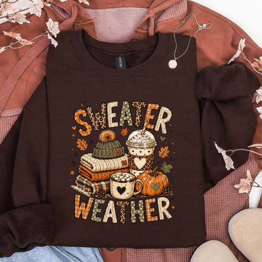 Brown sweatshirt featuring a Sweater Weather graphic with folded sweaters, knit hat, whipped coffee, cocoa mug, pumpkin, leaves, and leopard-print fall accents