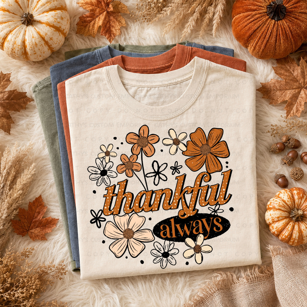 Folded cream Thanksgiving graphic T-shirt featuring orange, cream, and black retro flowers with the words Thankful Always, styled with pumpkins and autumn leaves.