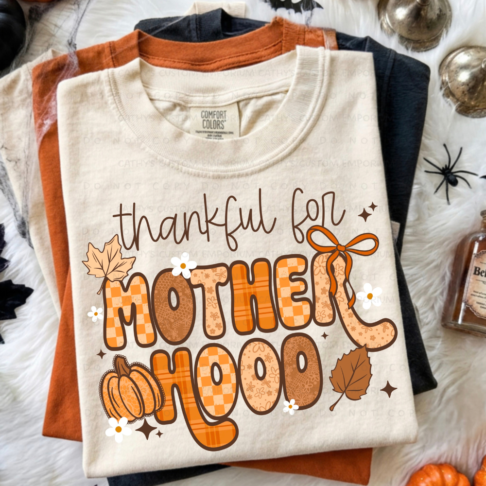 Folded cream Thanksgiving graphic T-shirt featuring Thankful for Motherhood lettering in orange, brown, plaid, and floral patterns with a pumpkin and autumn leaves.