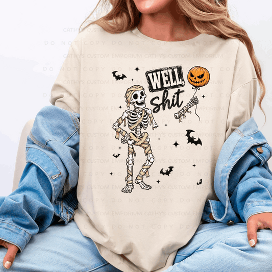 Cream sweatshirt featuring a wrapped mummy skeleton, flying bats, stars, a severed skeletal hand, and a jack-o-lantern balloon with the words Well Shit.
